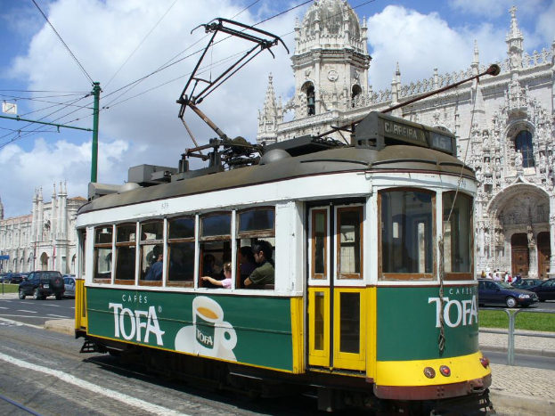 Eine Straßenbahn mit Menschen drinnen, Fahrzeuge auf der Straße, Gebäude, ein bewölkter Himmel, ein Strommast mit Drähten und ein Geländer in der Nähe.