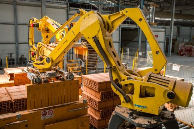 Ein großes Lager voller Kisten, Kräne und diverser industrieller Ausrüstung, mit einem Zaun und Pfählen im Vordergrund und einer Wand im Hintergrund.