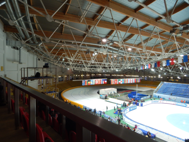 Großes Innen-Eisstadion mit zahlreichen Menschen auf dem Eis, umgeben von Sitzplätzen, Geländern, Brettern, Beleuchtungskörpern und konstruktiven Elementen, mit einer Wand im Hintergrund.