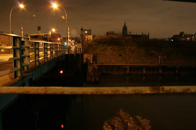Eine Brücke spannt einen Fluss mit zahlreichen Straßenlaternen darauf und im Hintergrund sind Gebäude zu sehen.