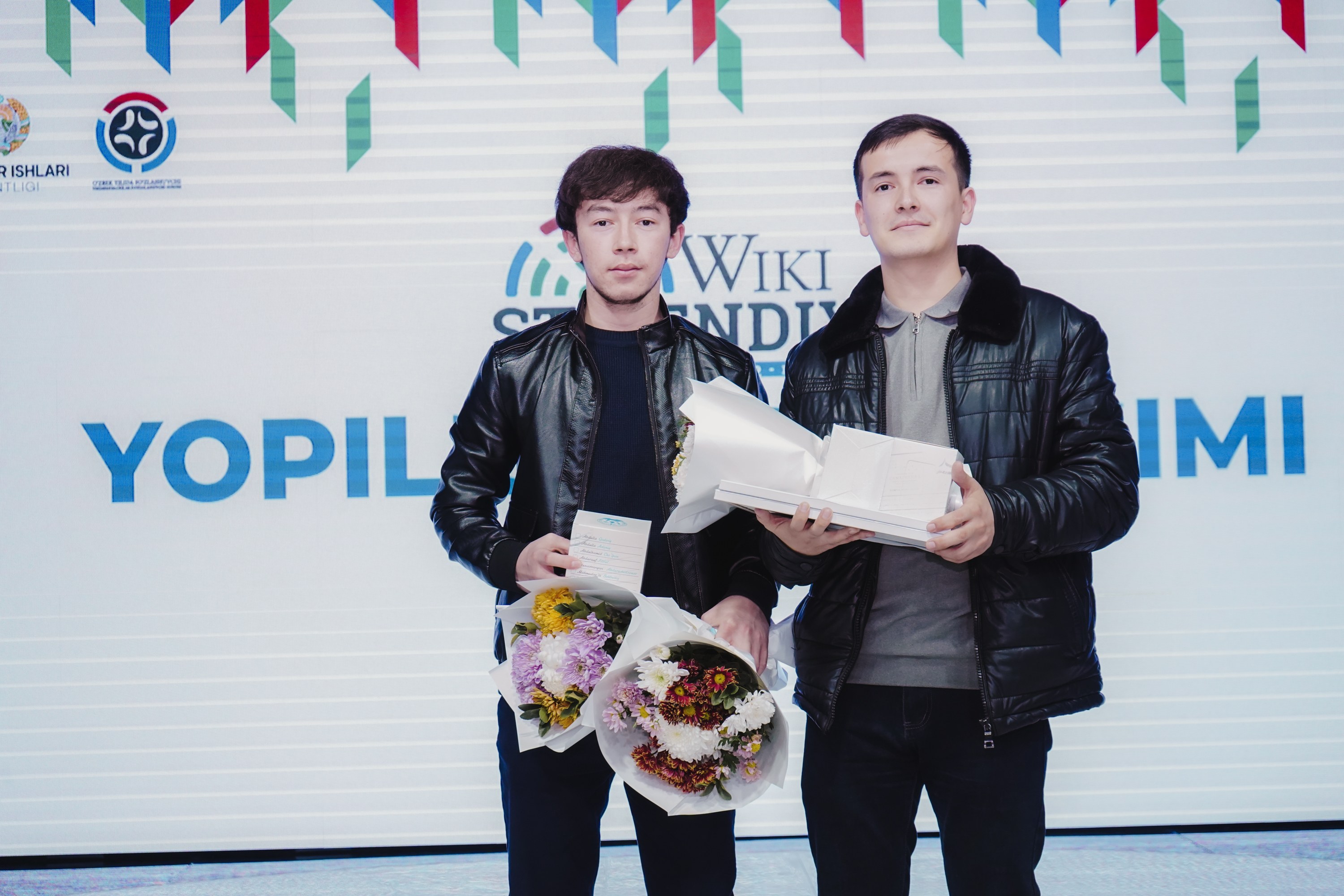 Zwei Männer in schwarzen Jacken stehen auf einer Bühne und halten Blumensträuße, im Hintergrund ein Banner des 'Yopil International Film Festival'.