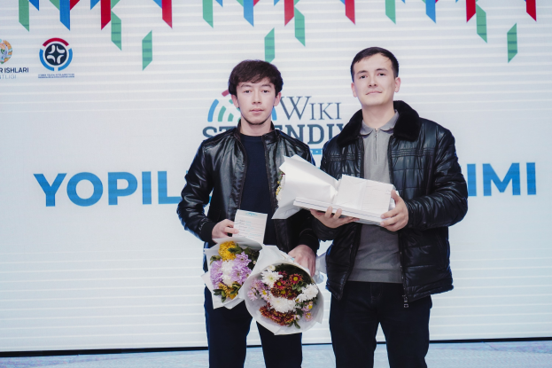 Zwei Männer in schwarzen Jacken stehen auf einer Bühne und halten Blumensträuße, im Hintergrund ein Banner des 'Yopil International Film Festival'.