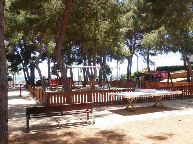 Eine Parkszene mit einem zentralen Tischtennis-Tisch, umgeben von Bänken, einem Zaun, Schaukeln, Pfählen, Pflanzen und Bäumen, mit einem Wasserlauf und einem klaren blauen Himmel im Hintergrund.