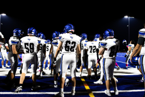 Eine Gruppe von Füßballspielern in Helmen und Stützschuhen steht auf einem Feld unter Flutlichtmästchen, bereit für ein Spiel.