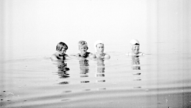 Vier Kinder schwimmen in einem Pool, alle tragen Schwimmkappen, dargestellt in Schwarz-Weiß.