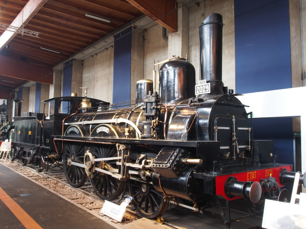 Alte Dampflokomotive in einem Museum ausgestellt, umgeben von Eisenbahnschienen, Informationsschildern, Ständen und anderen Ausstellungsstücken, mit einer Wand und Deckenbeleuchtung im Hintergrund.