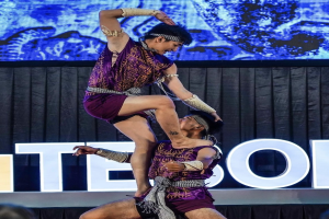 Zwei Performer in violetten Kleidern führen eine akrobatische Hebung auf der Bühne vor einem Publikum aus