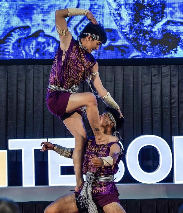 Zwei Performer in violetten Kleidern führen eine akrobatische Hebung auf der Bühne vor einem Publikum aus