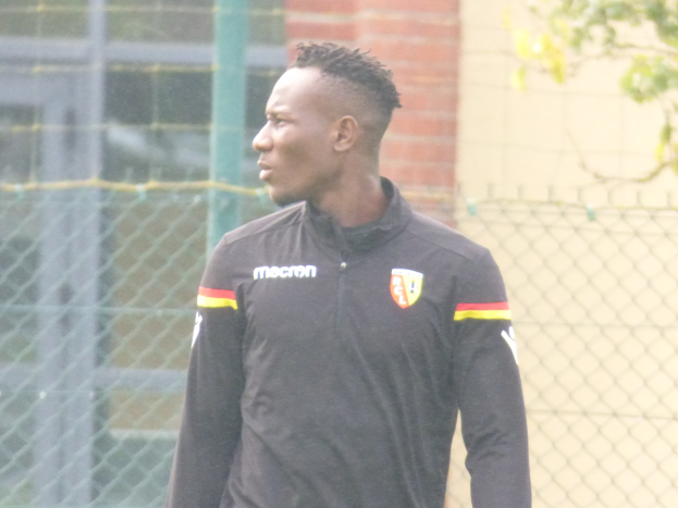 Ein Mann In Einer Schwarzen Jacke Steht Auf Einem Fussballfeld, Umgeben Von Einem Zaun Und Einem Baum, Scheinbar Fokussiert Während Einer Trainingseinheit.