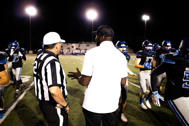 Ein Fußballtrainer mit Mütze spricht zu seinem Team auf einem Feld bei Nacht, mit Spielern in Helmen und Leuchten im Hintergrund unter einem dunklen Himmel.