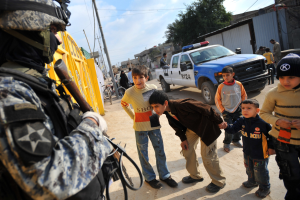 Eine Gruppe von Kindern vor einem Polizisten mit einer Waffe, mit Fahrzeugen, Menschen, Fahrrädern, Häusern, Bäumen, Mästen, Drähten und bewölktem Himmel im Hintergrund.