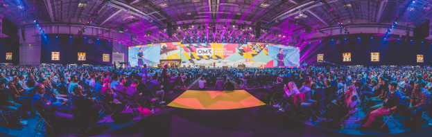 Eine große Menschenmenge sitzt auf Stühlen und schaut auf eine hell erleuchtete Bühne mit mehreren stehenden Personen, wahrscheinlich auf einem Online-Marketing-Rockstars-Event.