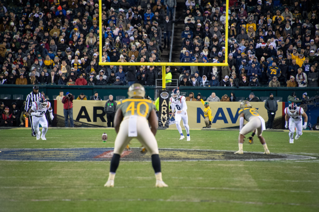 Fußballspieler in Sportkleidung und Helmen konkurrieren auf einem Feld mit Zuschauern im Hintergrund, Stadioninfrastruktur einschließlich Pfosten und Texttafeln sichtbar.