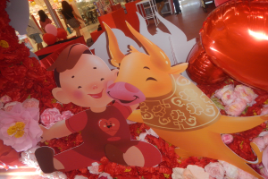 Eine festliche Szene mit Cartoon-Figuren auf einem mit Blumen und Ballons geschmückten Tisch, umgeben von Menschen, Stühlen, Tischen, Säulen, Lichtern und Geschäften, die ein chinesisches Neujahrsfest vermuten lassen.