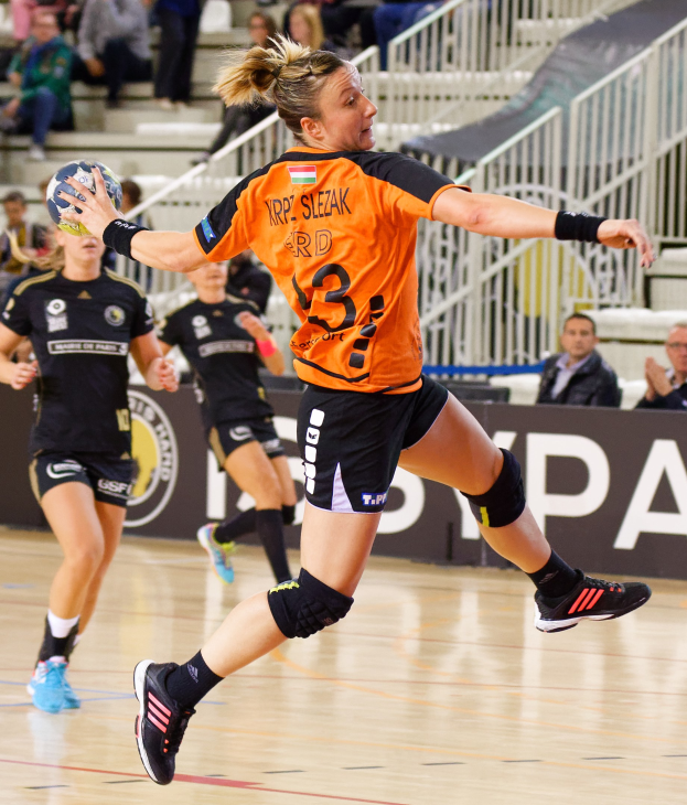 Eine Frau in einem orangen T-Shirt und schwarzen Shorts hält einen Handball, mit Zuschauern und einem Banner für die "Krp Slezak - Handball-Weltmeisterschaft 2019" im Hintergrund.