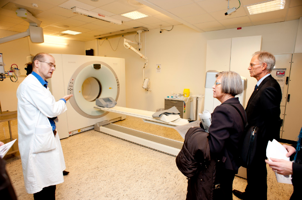 Ein Arzt in einem weißen Kittel erklärt eine CT-Scan-Maschine zwei Menschen in schwarzen Mänteln.