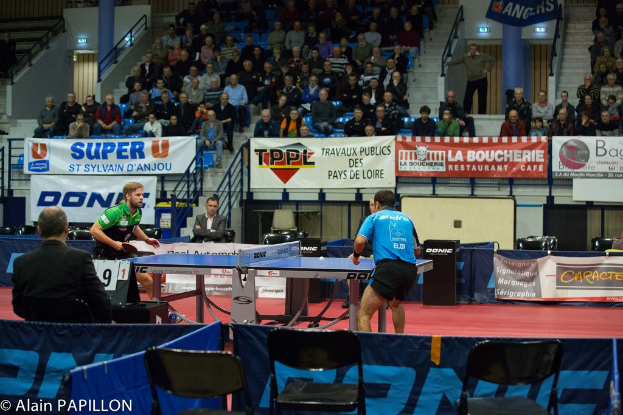 Zwei Männer spielen Tischtennis drinnen mit Zuschauern, die um die Tänzer stehen und sitzen, einige leere Stühle und Werbetafeln, mit Text in der unteren linken Ecke.