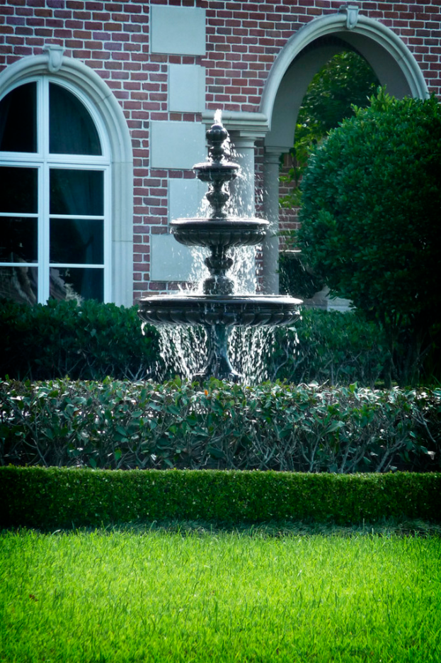 Eine GartenSzene mit Gras am Boden, einem zentralen Brunnen, umgeben von ein paar Pflanzen und einem Gebäude im Hintergrund.
