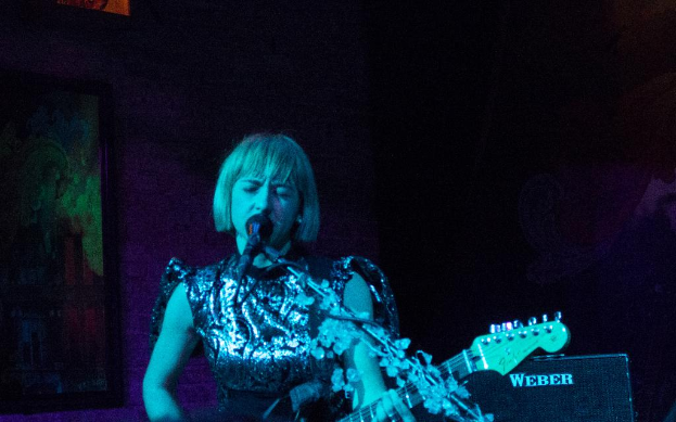 Eine Frau steht auf einer Bühne und singt in ein Mikrofon, während sie eine Gitarre spielt, mit einem Lautsprecher daneben und einer Wandmalerei im Hintergrund.