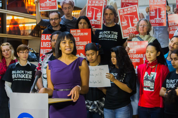 Eine Black-Lives-Matter-Aktivistin spricht in ein Mikrofon auf einem Podium und hält eine Tafel, während im Hintergrund eine Menge Menschen mit Schildern steht.