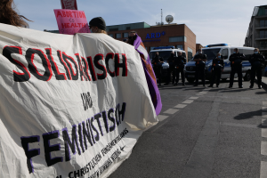 Eine Gruppe von Menschen marschiert auf einer Straße, hölt ein Banner mit der Aufschrift "Solidarität und Feminismus", mit parkenden Fahrzeugen am Rand und Gebäuden im Hintergrund unter einem klaren blauen Himmel.
