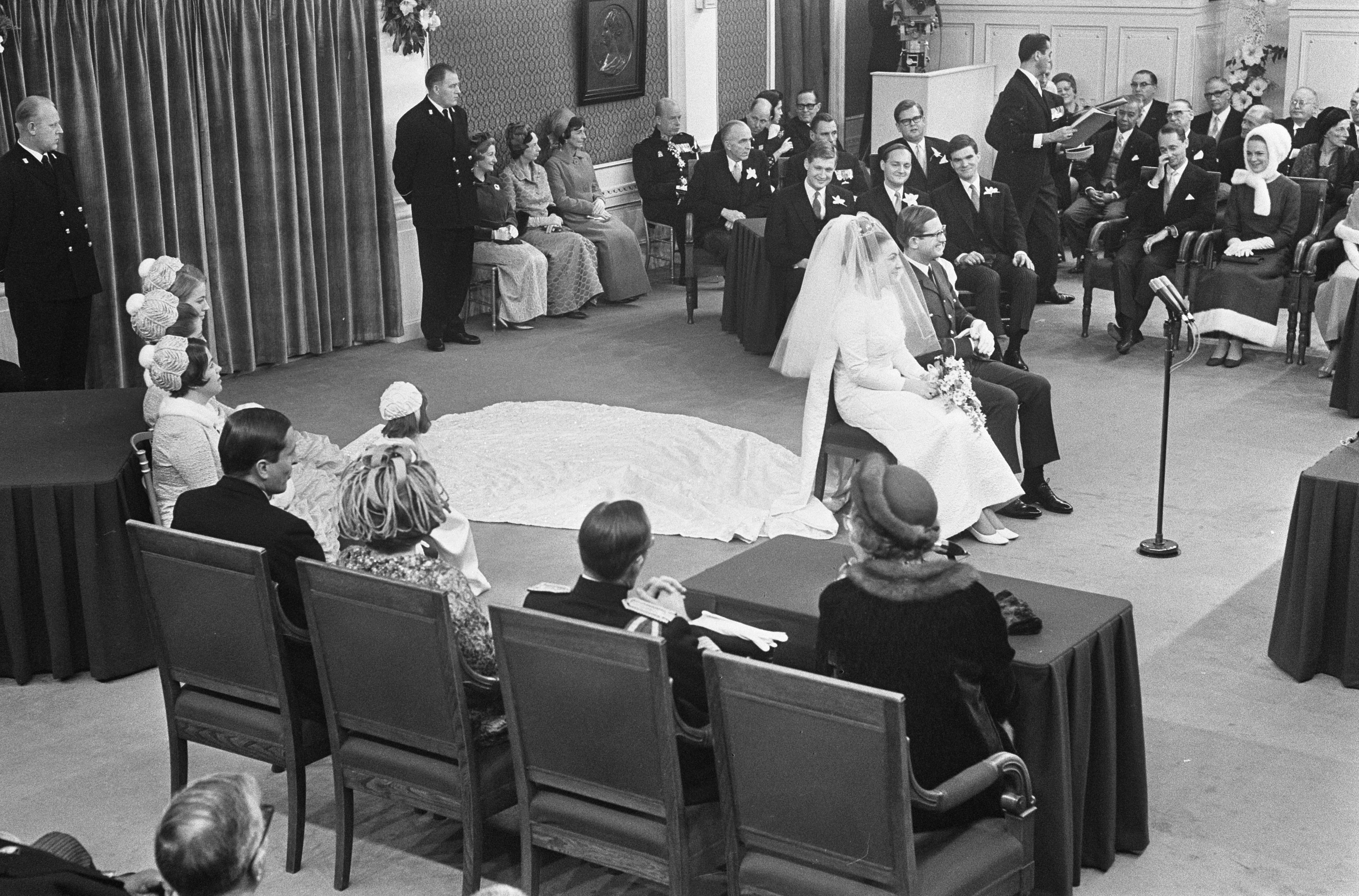 Schwarzes und weißes Foto einer Hochzeitszeremonie mit sitzenden und stehenden Gästen, einem Mann mit Mikrofon, einer Frau mit einem Bukett, einem Podium mit Mikrofon auf der rechten Seite und Blumenschmuck mit einem Vorhang auf der linken Seite.
