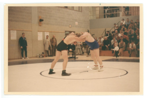 Zwei Wrestler kämpfen in der Mitte des Bildes, umgeben von Zuschauern, mit einer Wand voller angeklebter Papiere im Hintergrund.