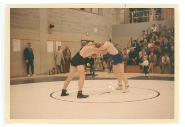 Zwei Wrestler kämpfen in der Mitte des Bildes, umgeben von Zuschauern, mit einer Wand voller angeklebter Papiere im Hintergrund.