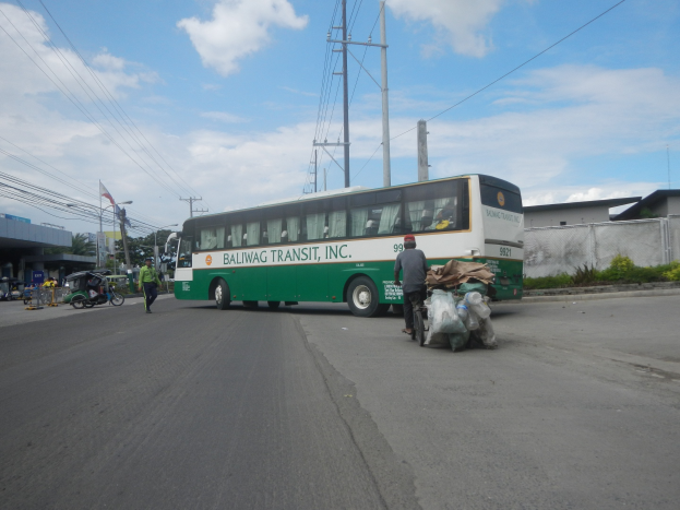 Ein grüner und weißer Bus fährt auf einer Straße neben einem Gebäude, mit Fußgängern und einem Fahrradfahrer in der Nähe, Strommasten, Gebäuden, Bäumen und einer bewölkten Himmel im Hintergrund.