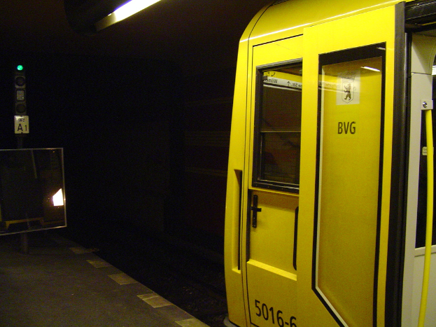 Ein Bahnhofsgebäude mit einem Zug auf der rechten Seite, einem Pfahl und einer Tafel auf dem linken Bahnsteig, einer Lampe oben und Bahnschienen unten.