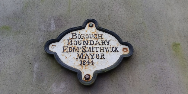 Schwarze Metalltafel an der Seite eines Gebäudes mit weißer, fetter, zentrierter Schrift, die "Gemeindegrenze Edm Smithwick Bürgermeister" liest.