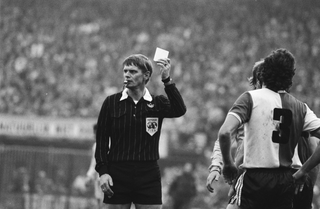 Ein Schiedsrichter in Schwarz-Weiß-Haltung hält eine rote Karte vor einer Menge von Menschen in Fußballkleidung und einem Zaun im Hintergrund.