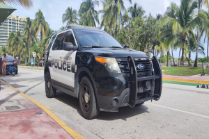 Polizeifahrzeug am Straßenrand mit Menschen, Fahrrädern, Gras, Bäumen, Gebäuden und einem klaren blauen Himmel im Hintergrund; das Vorderrad des Wagens ist sichtbar.