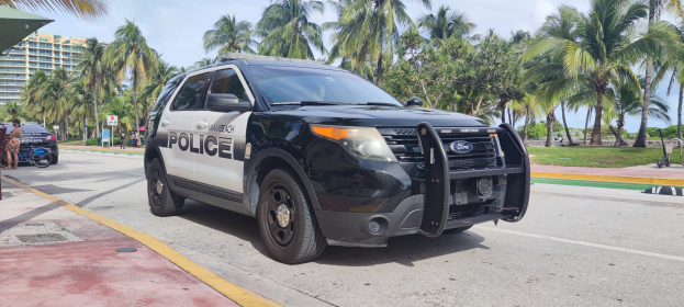 Polizeifahrzeug am Straßenrand mit Menschen, Fahrrädern, Gras, Bäumen, Gebäuden und einem klaren blauen Himmel im Hintergrund; das Vorderrad des Wagens ist sichtbar.