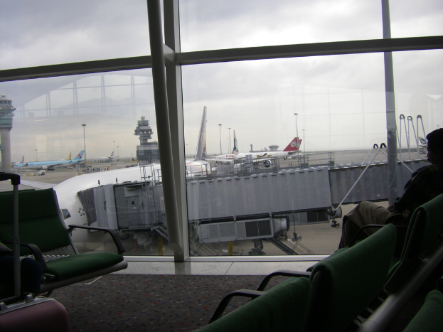 Ein Bild von hinter einem Fenster, das mehrere Stühle, einen Koffer auf der linken Seite und mehrere Flugzeuge und Abteile draußen zeigt.