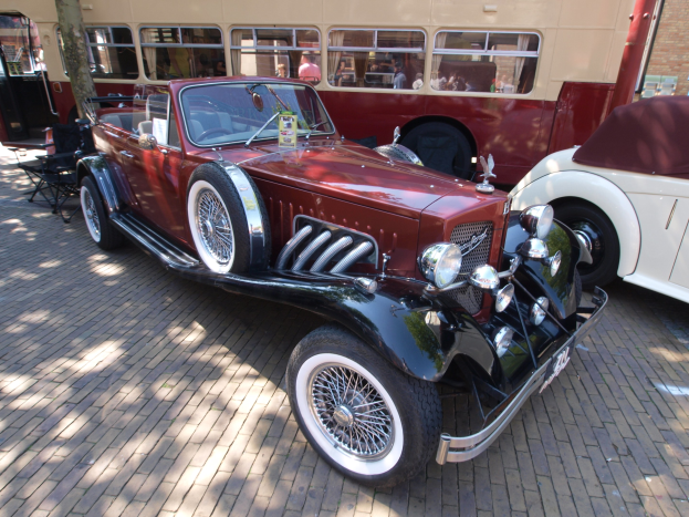 Vintage-Auto neben einem Doppeldeckerbus auf einer Kopfsteinpflasterstraße geparkt, mit Stühlen und einem Baum im Vordergrund und einem Gebäude im Hintergrund.