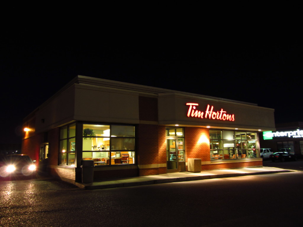 Ein Ladengeschäft mit der Aufschrift Tim Hortons, das Glasfenster und -türen hat und in dem Gegenstände auf Regalen zu sehen sind, neben einem fahrenden Auto mit Scheinwerfern, parkenden Autos in der Nähe und einem dunklen Himmel im Hintergrund.