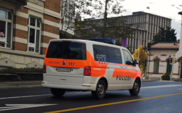 Ein Polizeiwagen fährt auf einer Straße mit hohen Gebäuden, Bäumen, Pflanzen, Pfählen, Verkehrszeichen und Schildern, mit dem Himmel im Hintergrund.