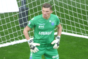 Ein Mann in einem grünen Torwarttrikot steht vor einem Fußballtor, trägt Handschuhe auf einem saftig grünen Rasen.