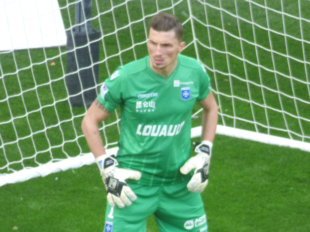 Ein Mann in einem grünen Torwarttrikot steht vor einem Fußballtor, trägt Handschuhe auf einem saftig grünen Rasen.