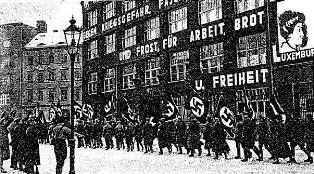 Schwarzes und wei├čes Bild einer Gruppe, die mit Fahnen und Spruchb├Ąndern die Stra├če entlangmarschiert, mit einem Lichtmast links und einer Tafel rechts mit der Aufschrift "Deutsche Nationalsozialistische Arbeiterpartei in Berlin".