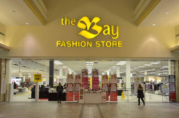 Außenansicht eines Bay Fashion Stores in einem Einkaufszentrum mit Menschen, Texttafeln an Wänden, Säulen, Deckenleuchten und Hintergrundelementen.