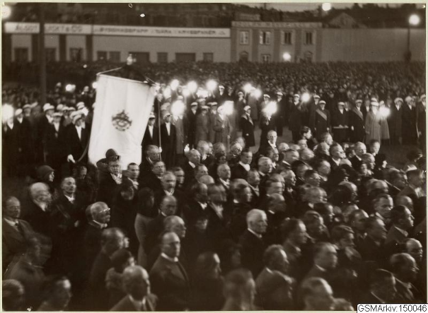 Schwarzes und weißes Foto einer großen Menschenmenge vor einem Gebäude, mit einer Person, die eine Fahne hält, umgeben von Pfosten und Lichtern; Text unten lautet "Gustav Klimts Rede an der Berliner Mauer."