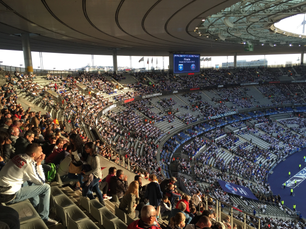 Große Menschenmenge in einem Stadion bei einem Fußballspiel, mit einer Bühne rechts, Fahnen, Stangen, einem Bildschirm und dem Allianz Stadion in München, Deutschland im Hintergrund.