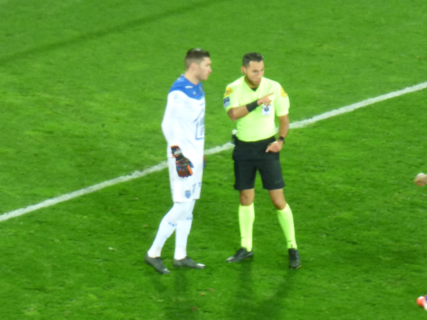 Ein Schiedsrichter in einem gelben T-Shirt und schwarzen Shorts spricht mit einem Fußballspieler in einer weißen und blauen Uniform auf dem Feld, beide tragen Turnschuhe.