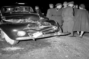 Ein Schwarz-Weiß-Bild eines verunglückten Autos am Straßenrand mit eingedellter Motorhaube und zersplitterten Fenstern, umgeben von einer Gruppe besorgter Menschen, die den Schaden begutachten.