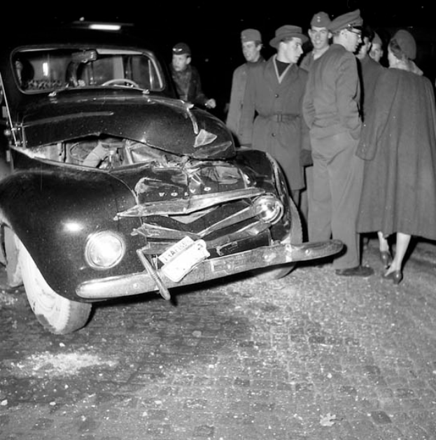 Ein Schwarz-Weiß-Bild eines verunglückten Autos am Straßenrand mit eingedellter Motorhaube und zersplitterten Fenstern, umgeben von einer Gruppe besorgter Menschen, die den Schaden begutachten.