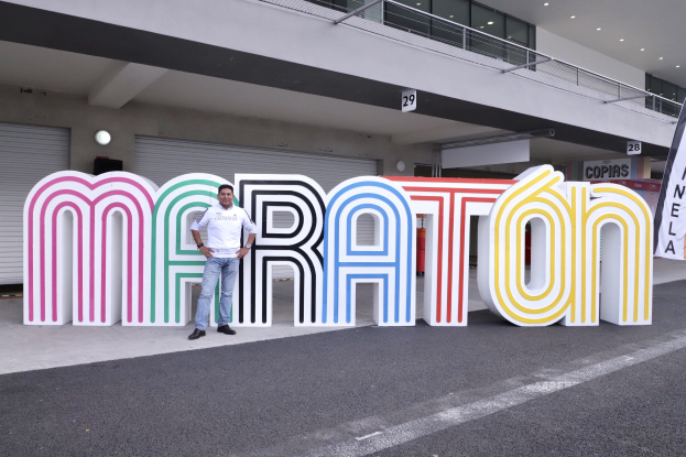 Ein Mann steht am Start eines Marathons vor einem großen "Marathon"-Schild mit einem Banner rechts daneben, im Hintergrund ein Gebäude mit Lichtern, Geländern und Fensterläden.