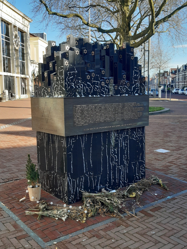 Denkmal in einem Stadtplatz für Holocaust-Opfer mit einem Baum davor, einem Topf mit einer Pflanze auf dem Boden, verstreuten trockenen Blättern und Gebäuden, Fahrzeugen, Masten und Bäumen im Hintergrund.
