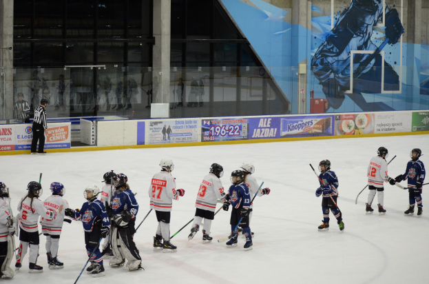 Junge Menschen in Helmen und Eishockeyausrüstung spielen Eishockey auf einer Indoor-Eisbahn mit Bannern, Säulen und Glaswänden im Hintergrund.